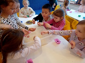 Dzieci siedzą przy stole i dotykają rękami kawałków gliceryny, którą wyciągają z pudełka.