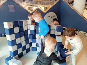 5 dzieci buduje dom z białych i granatowych kwadratowych klocków. Jeden chłopiec stoi w środku i się śmieje a pozostałe dzieci budują ściany. 