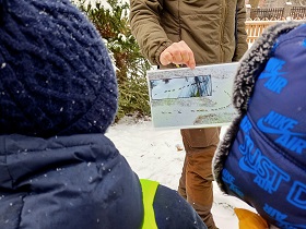 Pan w oliwkowym stroju pokazuje dwójce dzieci zdjęcie, na którym widać watahę wilków idącą po śniegu.