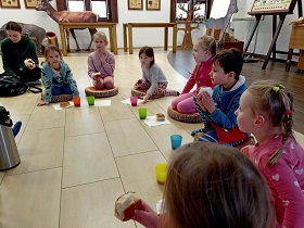 Dzieci siedzą w sali na okrągłych poduszkach i jedzą bułki. Przed nimi znajdują się kolorowe kubki z piciem. 