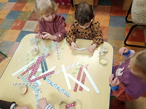 4 dziewczynki siedzą przy stole i tworzą łańcuch choinkowy z kolorowych pasków papieru. Na stole leży dużo wyciętych kolorowych papierowych pasków. 