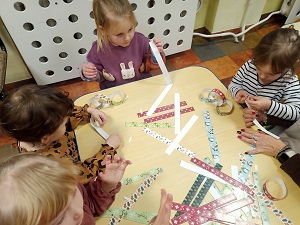 4 dziewczynki siedzą przy stole i tworzą łańcuch choinkowy z kolorowych pasków papieru. Na stole leży dużo wyciętych kolorowych papierowych pasków. 