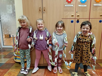 4 śmiejące się dziewczynki stoją w sali i mają założony na siebie łańcuch choinkowy wykonany z papierowych pasków. 