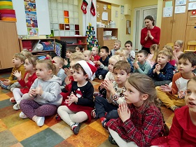 Dzieci siedzą w sali na podłodze, klaszczą w ręce i słuchają zimowej piosenki. 