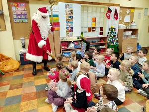 Dzieci siedzą w sali na podłodze, klaszczą w ręce i słuchają zimowej piosenki. Przed dzieci wchodzi Mikołaj, który wota się uściskiem dłoni z jednym z chłopców.