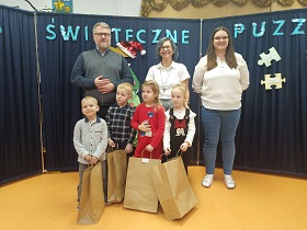 Czworo dzieci oraz dwie panie i pan stoją i pozują do zdjęcia. Za nimi znajduje się granatowy parawan z napisem konkurs świąteczne puzzle. Dzieci trzymają w dłoniach brązowe torby.