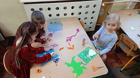 Trzy dziewczynki siedzą przy stoliku i tworzą kartki świąteczne dla dzieci z Domu Dziecka. Używają do tego kartek, bibuły i naklejek. Na kartkach widać bałwanki i choinkę z ruchomymi i sensorycznymi elementami.