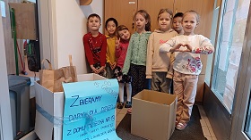 Siedmioro dzieci stoi przy dwóch pudłach z darami dla Domu Dziecka. Na jednym z pudełek przywiązany jest plakat informacyjny o akcji.