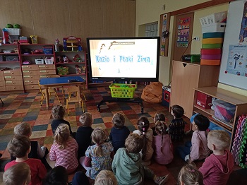 Dzieci siedzą tyłem do zdjęcia, w sali na podłodze. Przed nimi stoi tablica multimedialna na ekranie której wyświetlony jest film Kazio i ptaki zimą. 