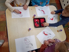 5 dzieci siedzi przy stole. Przed nimi leżą kartki z szablonami gila. Na środku stołu stoi pojemnik z kawałkami czerwonej bibuły i kleje. Dzieci przyklejają czerwona bibułę na brzuszek ptaszka. 