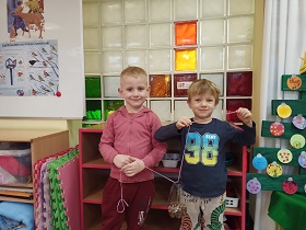 Dwóch chłopców stoi obok siebie w sali. W rękach trzymają sznurki, na których zawieszony jest karmnik z ziarenkami wykonany z plastikowej butelki. 