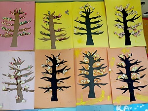 Na stoliku znajduje się 8 prac plastycznych, przedstawiających drzewa w różnych porach roku na kolorowych kartkach. Drzewa mają przyczepiony popcorn, który jest pomalowany farbami. 