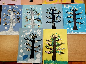 Na stoliku znajduje się 6 prac plastycznych, przedstawiających drzewa w różnych porach roku na kolorowych kartkach. Drzewa mają przyczepiony popcorn, który jest pomalowany farbami. 