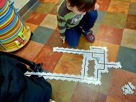 Chłopiec w bluzce w paski siedzi na podłodze i spogląda na ułożone drewniane puzzle, na których znajduję się mały robocik - ozobot. 
