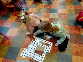 Dwie dziewczynki siedzą na podłodze i układają drewniane puzzle. 