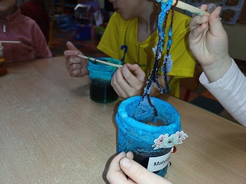 Przy stoliku siedzą dzieci i obserwują swoje wyhodowane kryształy osadzone na sznurkach włóczki przywiązanych do patyczka. Dwa słoiki zawierają roztwór soli zabarwionej na niebiesko. 