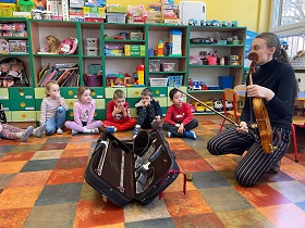 Dzieci siedzą w kole na podłodze. Na środku leży futerał na instrument. Pani trzyma altówkę ze smyczkiem i opowiada dzieciom o tym instrumencie. 
