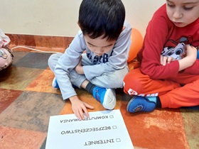 Dwóch chłopców, jeden ubrany na szaro, drugi na czerwono, siedzą na podłodze i czytają kartkę z napisem: Internet, Bezpieczeństwo, Komputeromania