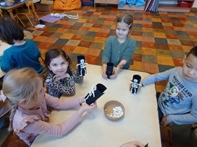 Czworo dzieci siedzi przy stoliku. Prezentują wykonane szkielety ze słomek, przyklejone do czarnej rolki z brystolu
