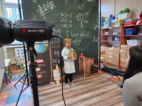 Chłopiec trzyma zabawkowe fiolki chemiczne przebrany za naukowca. Pani fotograf robi mu zdjęcia na tle kotary obrazującej tablicę w szkole z różnymi równaniami matematycznymi, chemicznymi itp.
