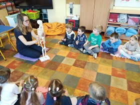 Dzieci siedzą na podłodze na matach. Pani Karolina siedzi razem z dziećmi w kole i prezentuje szkielet człowieka.