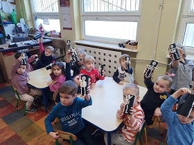 Dzieci siedzą przy stoliku. Prezentują wykonane szkielety ze słomek, przyklejone do czarnej rolki z brystolu.