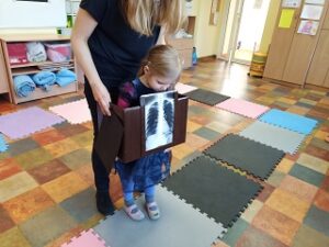 Pani Karolina trzyma brązowe pudełko z przyklejonym zdjęciem rentgenowskim klatki piersiowej. W pudełku stoi dziewczynka w granatowej sukience, imitując, że jest to jej zdjęcie.