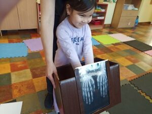 Pani Karolina trzyma brązowe pudełko z przyklejonym zdjęciem rentgenowskim dłoni. Do pudełka wkłada ręce dziewczynka w fioletowej bluzce, imitując, że jest to zdjęcie jej dłoni.