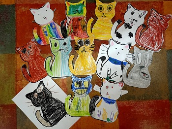 Prace plastyczne dzieci leżą na podłodze. Są to prace przedstawiające koty. Koty są pomalowane pastelami. Każdy kot wygląda inaczej i ma inny kolor. 