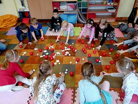 Dzieci schylają się by podnieść czerwone plastikowe piłeczki ze środka kółka. Na środku podłogi znajdują się również białe kulki oraz białe gwiazdki. 