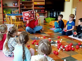 Dzieci siedzą w półkolu i obserwują panią, która pokazuje im obrazek, na którym przedstawione są czerwone i białe krwinki oraz płytki krwi. 