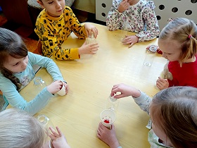 Szóstka dzieci siedzi przy stoliku i wkłada czerwone i białe kulki do plastikowej butelki.