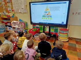 Dzieci siedzą na podłodze i oglądają obrazek wyświetlany na monitorze, który przedstawia piramidę zdrowia. 