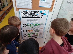 Dzieci stoją przy plakacie ,,Jakie jest dziś powietrze w Warszawie?”. Za pomocą magnesu będą zaznaczać jaką jakość powietrza mieli tego dnia. 
