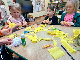 Trzy dziewczynki siedzą przy stoliku i robią z krepiny żółte żonkile. Na stoliku znajduje się biały sznurek, żółta i pomarańczowa krepina, nożyczki i klej.