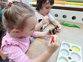 Dziewczynka i chłopiec przy stoliku malują farbami szyszki na żółto.