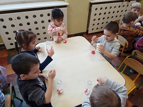 3 chłopców i 2 dziewczynki siedzą przy stoliku. Każde z dzieci trzyma w ręku przezroczystą, plastikową butelkę z wsypanymi czerwonymi i białymi pomponami. Dziewczynka i chłopiec trzymają również białe kubki, z których przesypują pompony. 