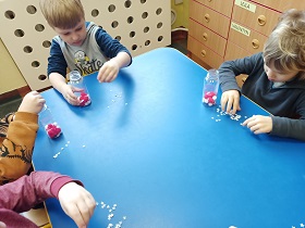 4 dzieci siedzi przy stoliku. Każde z dzieci trzyma w ręku plastikową butelkę wypełniona białymi i czerwonymi pomponami. Obok butelek leżą papierowe, błyszczące gwiazdki, które dzieci wrzucają do butelek. 