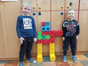 2 chłopców stoi obok wysokiego robota wykonanego z dużych, kolorowych, plastikowych klocków wafle. 