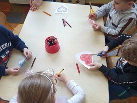 4 dzieci siedzi przy stole. Każde z dzieci ma przed sobą pokolorowane serce. Jeden chłopiec wycina serce nożyczkami. 
