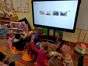 Dzieci siedzą przed monitorem, na którym wyświetlana jest prezentacja o pojazdach. Dzieci obserwują zdjęcia pojazdów ustawionych w rzędzie. Część dzieci siedząca w pierwszym rzędzie podnosi rękę. 