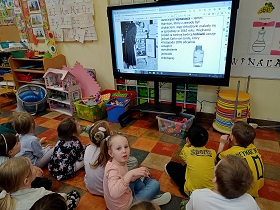 Dzieci siedzą przed monitorem, na którym wyświetlana jest prezentacja o wynalazkach. Dzieci spoglądają na zdjęcie pani przy starej lodówce oraz na napisy przy zdjęciu. Jedna dziewczynka w różowej bluzce patrzy na panią. 