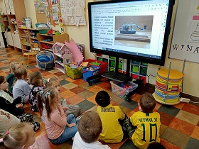 Dzieci siedzą przed monitorem, na którym wyświetlana jest prezentacja o wynalazkach. Dzieci spoglądają na zdjęcie telegramu i napisy przy zdjęciu.