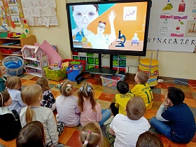 Dzieci siedzą przed monitorem, na którym wyświetlana jest piosenka o wynalazkach. Na filmie widać panią w okularach oraz stojące z a nią maszyny do robienia eksperymentów. 