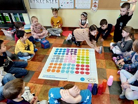 Dzieci siedzą na podłodze na poduszkach wokół maty do kodowania. Dziewczynka w beżowej bluzie i kucykach sięga po szary kubeczek, by móc go postawić na macie na odpowiednim koloru planszy. 