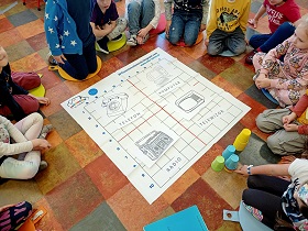 Dzieci siedzą na podłodze na poduszkach wokół maty do kodowania. Na planszy znajdują się cztery rysunki i napisy - telefon, radio, komputer i telewizor.