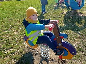 Chłopiec w żółtej czapce i odblaskowej kamizelce siedzi na zabawkowym motorku. 