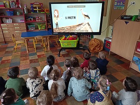Dzieci siedzą na podłodze przed tablicą multimedialną. Na ekranie wyświetlony jest film edukacyjny o bocianach. 