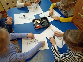 5 dzieci siedzi przy stole i wykleja szablony bocianów przy pomocy plasteliny. Pośrodku leży pudełko z kawałkami białej, czarnej, szarej i czerwonej plasteliny. 