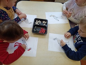 4 dzieci siedzi przy stole i wykleja szablony bocianów przy pomocy plasteliny. Pośrodku leży pudełko z kawałkami białej, czarnej, szarej i czerwonej plasteliny. 